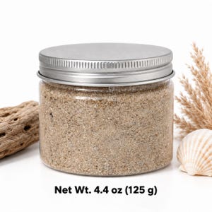 Closed clear jar of deep teal green colored quartz sand with a silver metal screw lid, next to a monstera leaf, a white ceramic pineapple with a gold top and two cowrie shells, with text: Net Wt.