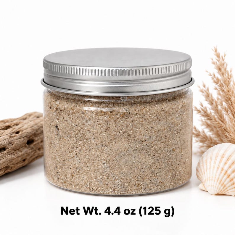 Closed clear jar of deep teal green colored quartz sand with a silver metal screw lid, next to a monstera leaf, a white ceramic pineapple with a gold top and two cowrie shells, with text: Net Wt.