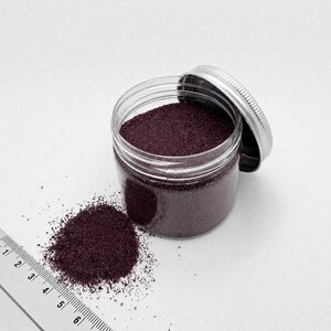 Open jar of Plum Velvet plum purple quartz sand with its silver lid, next to a small pile of spilled sand and a ruler in centimeters showing the fine grain size