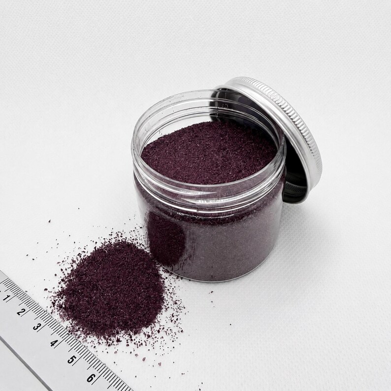 Open jar of Plum Velvet plum purple quartz sand with its silver lid, next to a small pile of spilled sand and a ruler in centimeters showing the fine grain size