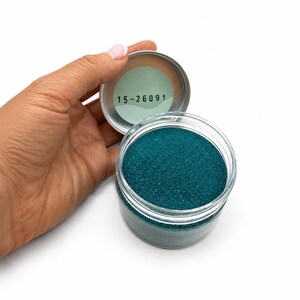 Top view of an open jar of Ocean Breeze teal quartz sand, with a hand holding the silver lid, which has a label with the number 15-26091 on the inside