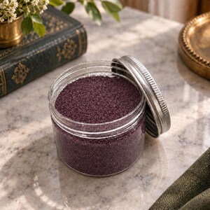 Open jar of Plum Velvet plum purple quartz sand with its silver lid leaning against it, on a marble surface next to an old green book, a small brass vase with white flowers and a brass tray