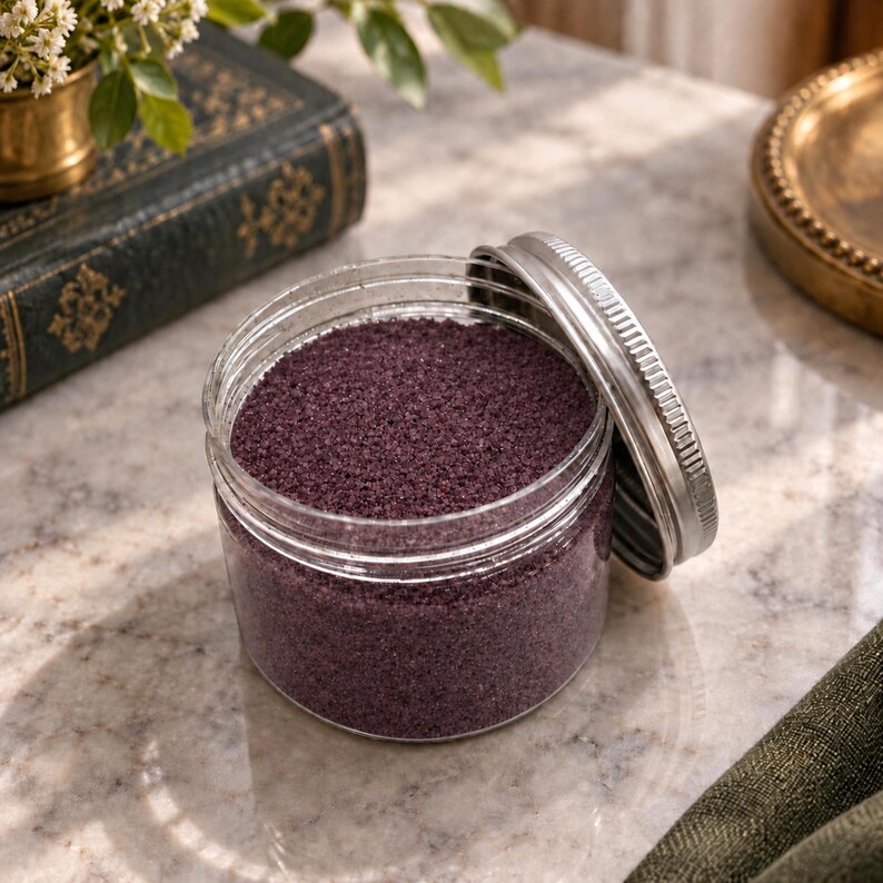 Open jar of Plum Velvet plum purple quartz sand with its silver lid leaning against it, on a marble surface next to an old green book, a small brass vase with white flowers and a brass tray