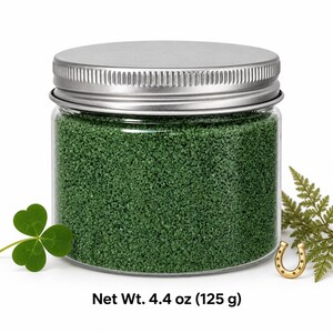 Closed clear jar of forest green colored quartz sand with a silver metal screw lid, next to a three-leaf clover, a small gold horseshoe and a fern sprig, with text: Net Wt. 4.4 oz (125 g).