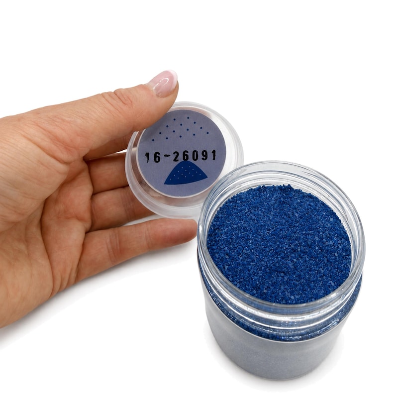 Top view of an open jar of Ocean Depth deep blue quartz sand, with a hand holding the lid, which has a label with the number 16-26091 and a small sand pile illustration on the inside