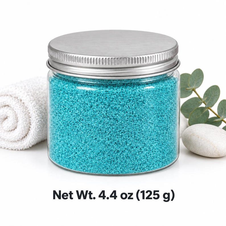 Closed clear jar of turquoise colored quartz sand with a silver metal screw lid, next to a rolled white towel, a eucalyptus sprig and a smooth white stone, with text: Net Wt. 4.4 oz (125 g).