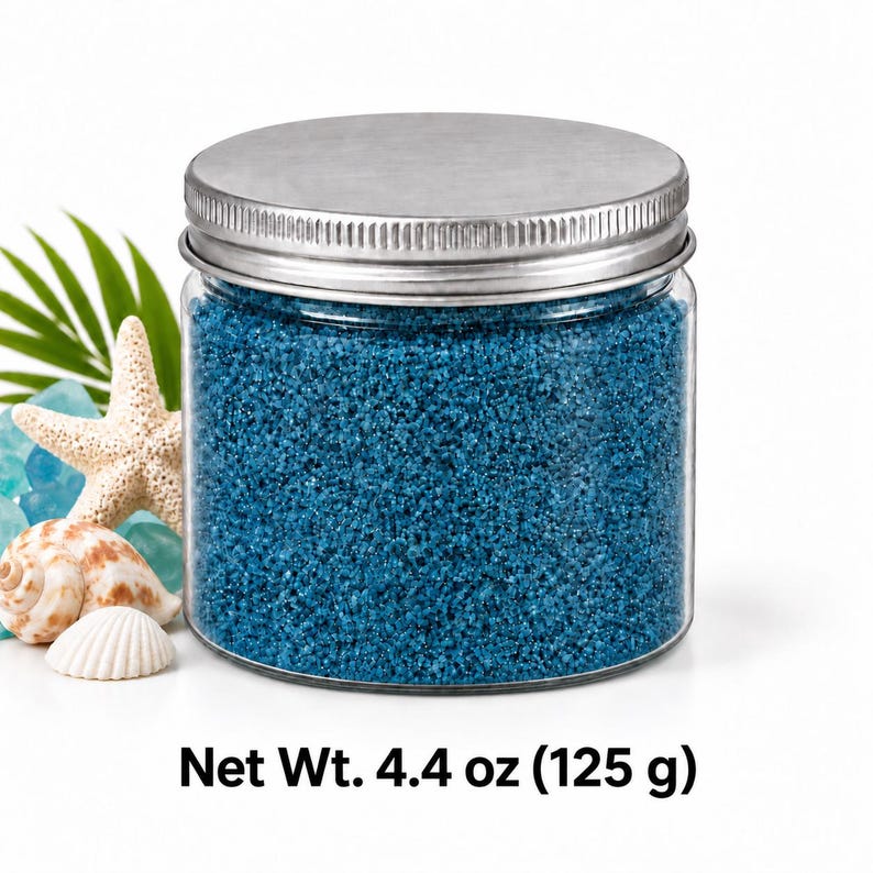 Closed clear jar of ocean blue colored quartz sand with a silver metal screw lid, next to a starfish, seashells, sea glass and a palm leaf, with text: Net Wt. 4.4 oz (125 g).
