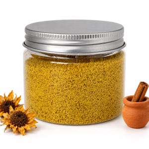 Closed clear jar of Golden Saffron colored quartz sand in a mustard yellow, with a silver metal screw lid, next to two dried sunflower heads and a small terracotta pot holding cinnamon sticks, on a white background.