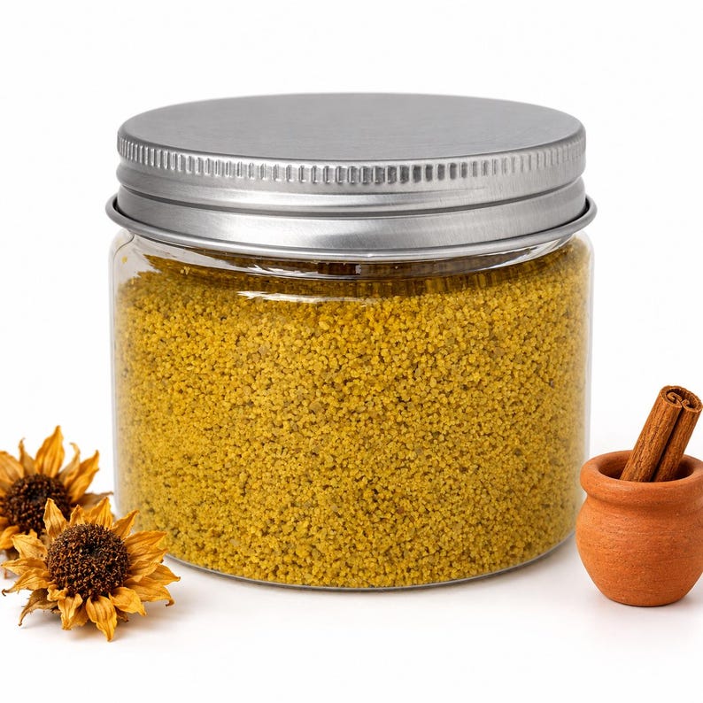 Closed clear jar of Golden Saffron colored quartz sand in a mustard yellow, with a silver metal screw lid, next to two dried sunflower heads and a small terracotta pot holding cinnamon sticks, on a white background.