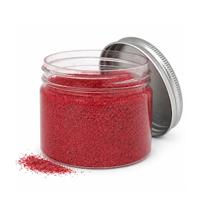 Open clear jar of deep red colored quartz sand with its silver metal screw lid leaning behind it and a small pile of the same sand spilled in front, showing the fine grain texture, on a white background.