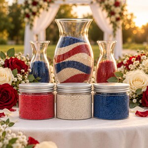 Three closed clear jars of red, off-white and royal blue quartz sand in front of a glass ceremony vase with wavy red, white and blue layers. Two smaller vases hold the blue and red sand. Red and white roses and baby’s breath surround them, with a white wedding arch at sunset behind.