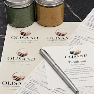 K&ouml;nnte beinhalten: Zwei durchsichtige Gl&auml;ser mit gr&uuml;nem und beigem Quarzsand stehen auf einem Bogen mit Olisand-Etiketten und einer Dankeskarte. Ein silberner Kugelschreiber liegt diagonal auf dem Papier. Der Text lautet Olisand Handcrafted Colored Quartz Sand.