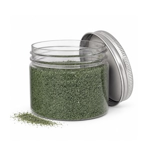 Open clear jar of muted olive green colored quartz sand with its silver metal screw lid leaning behind it and a small pile of the same sand spilled in front, showing the fine grain texture, on a white background.