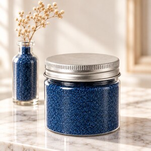 Closed clear jar of royal blue colored quartz sand with a silver metal screw lid on a white marble surface in soft sunlight, next to a small glass bottle of the same blue sand holding dried flowers.