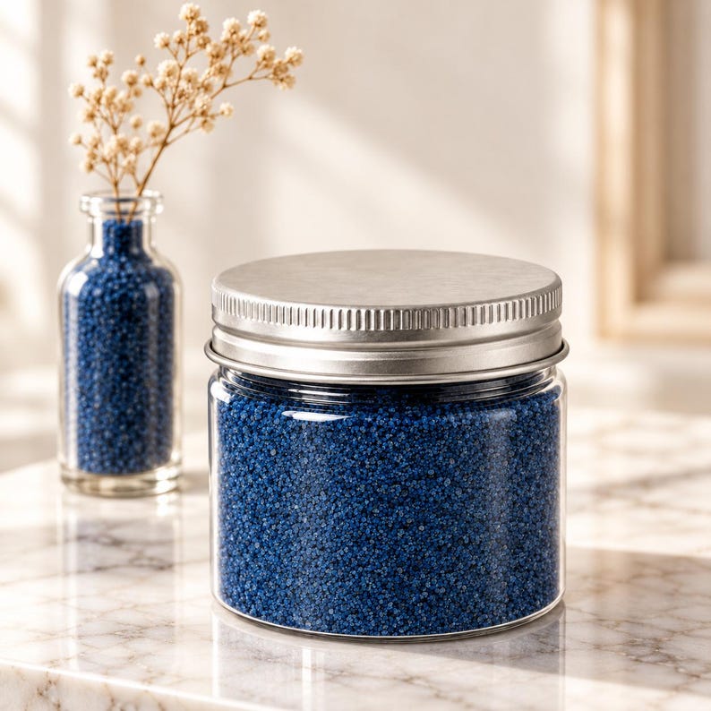 Closed clear jar of royal blue colored quartz sand with a silver metal screw lid on a white marble surface in soft sunlight, next to a small glass bottle of the same blue sand holding dried flowers.