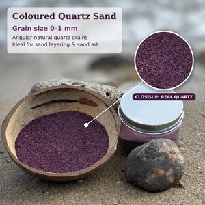 Coconut shell bowl filled with Plum Velvet colored quartz sand in a deep plum purple, next to a closed jar of the same sand with a silver metal screw lid, a grey stone and a piece of coral on a beach. A magnified circle shows the fine angular grains, labeled Close-up: Real Quartz. Text: Coloured Quartz Sand, grain size 0-1 mm, angular natural quartz grains, ideal for sand layering and sand art.