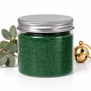 Closed clear jar of deep green colored quartz sand with a silver metal screw lid, next to a eucalyptus sprig and a gold jingle bell with a star cutout on a gold cord, on a white background.