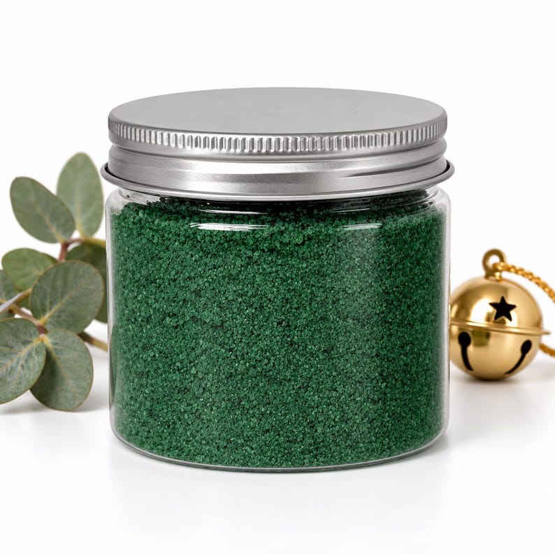 Closed clear jar of deep green colored quartz sand with a silver metal screw lid, next to a eucalyptus sprig and a gold jingle bell with a star cutout on a gold cord, on a white background.