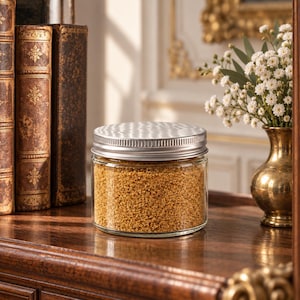 Closed clear jar of Amber Gold colored quartz sand in a warm golden amber, with a silver metal screw lid, on a polished wooden shelf next to antique leather-bound books and a brass vase of small white flowers, in an elegant room with gilded details.