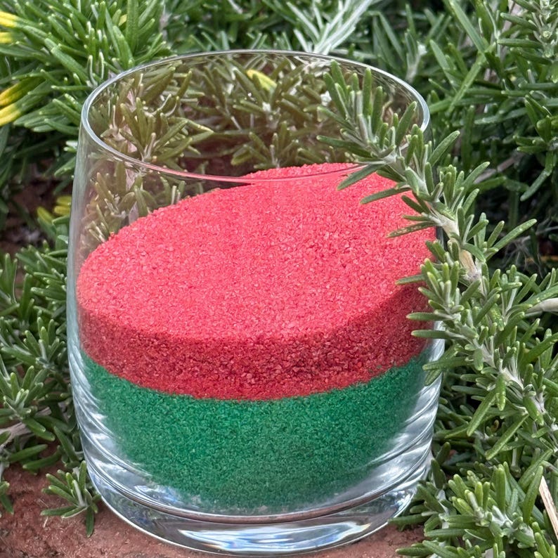 Glass tumbler filled with layered Canyon Rose red sand and green sand, photographed outdoors among rosemary greenery