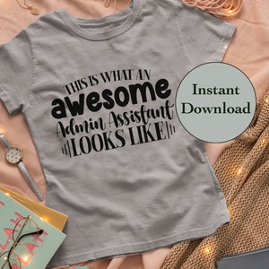May include: A gray t-shirt with the text "This is what an awesome admin assistant looks like" printed in black. The t-shirt is lying on a pink and brown blanket with string lights.