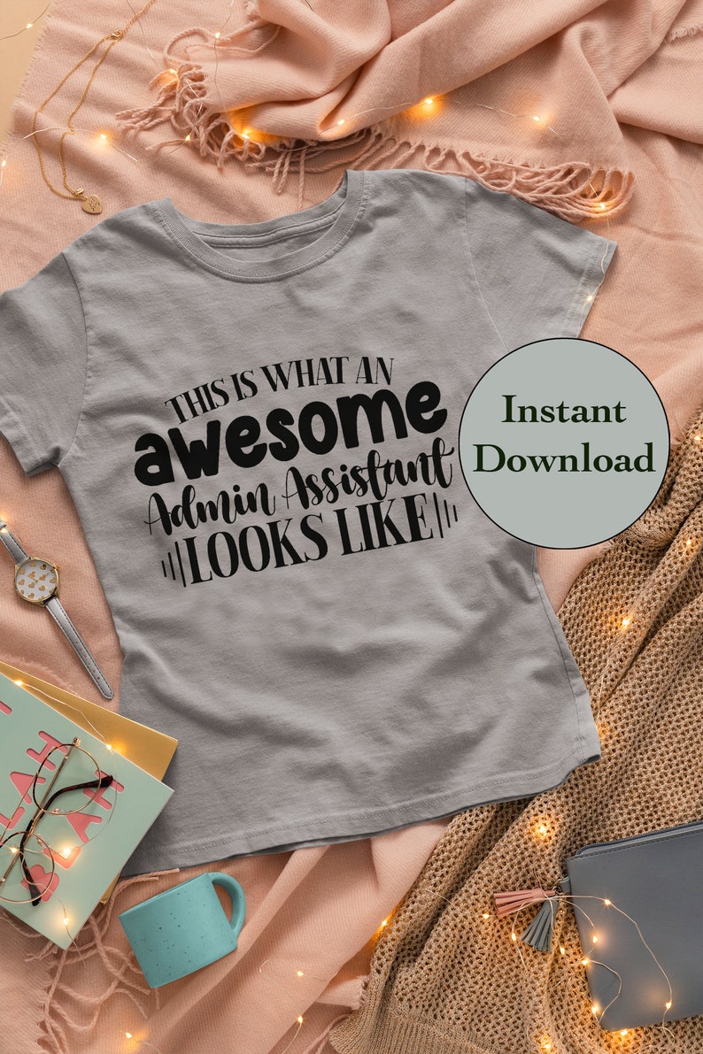 May include: A gray t-shirt with the text "This is what an awesome admin assistant looks like" printed in black. The t-shirt is lying on a pink and brown blanket with string lights.