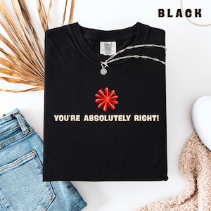 Könnte beinhalten: Ein schwarzes T-Shirt mit einer roten Sternengrafik und dem Text "YOU'RE ABSOLUTELY RIGHT!" in heller Schrift. Das T-Shirt hat ein Comfort Colors Markenetikett am inneren Halsausschnitt und wird mit einer silbernen Halskette präsentiert.