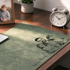 Puede incluir: Un protector de escritorio de color verde salvia con una ilustración grabada personalizada de un hombre con dos niños y el texto "We love Daddy" en la esquina inferior, sobre un escritorio de madera junto a un portátil.