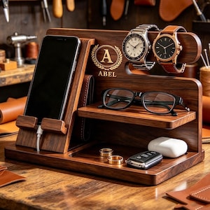 Puede incluir: Una estación de carga de madera oscura con soporte para smartphone y cable, un estante para gafas y una barra horizontal para dos relojes de pulsera. La madera está grabada con un emblema de corona de laurel, la letra A y el nombre ABEL.