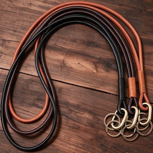 May include: Three long, rounded leather cords in black, dark brown, and tan. Each cord features a wrapped leather detail at the end, connected to a brass-toned metal oval carabiner and a circular split ring for holding keys or other small items.