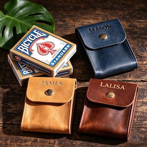 May include: Three leather card cases in navy, tan, and dark brown, each featuring a brass snap closure and a personalized name embossed on the flap. Next to them are two stacked Bicycle Standard playing card decks with blue and white packaging.