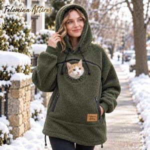 May include: An olive green sherpa fleece hoodie featuring a large front pouch with a circular opening containing an orange and white cat. The hoodie includes a hood with drawstrings, long sleeves with ribbed cuffs, and a leather-style patch with the text Felomina Attires.
