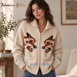 May include: A cream-colored knit zip-up cardigan featuring a shawl collar and two front pockets. The garment displays two fox head graphics in brown, black, and red, with light blue paw print patterns near the ribbed cuffs and hem.
