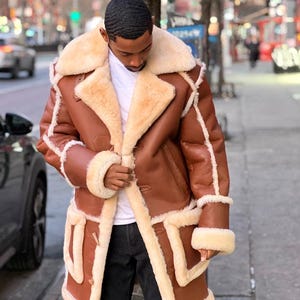 May include: A brown leather coat with a thick cream-colored shearling lining, featuring a large fold-over collar, long sleeves with shearling cuffs, and two large patch pockets on the front. The coat is worn over a white shirt and black pants.