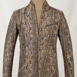 May include: A blazer featuring a brown, black, and tan snakeskin pattern. The garment has a mandarin collar, a single button closure at the front, and a horizontal zippered pocket on the upper left chest area, displayed on a mannequin.