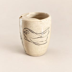 May include: A cylindrical ceramic cup with a speckled cream-colored glaze. The exterior features hand-drawn black line art of two ghosts. The rim of the cup has a small notch cut out on one side, and the surface has a smooth, glossy finish.