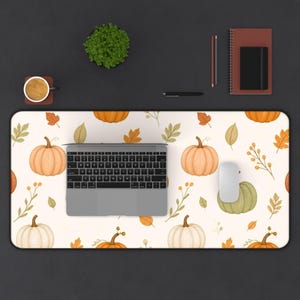 Op de afbeelding: Een rechthoekige bureaumat met een herhalend patroon van oranje en groene pompoenen, herfstbladeren en takjes op een crèmekleurige achtergrond. Een zilveren laptop, een witte computermuis, een mok, een plantje en een notitieboek liggen erop.