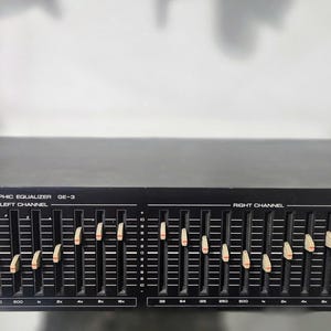 May include: A black Yamaha Natural Sound Stereo Graphic Equalizer GE-3 featuring a 10-band dual channel system. The front panel includes a power button with a red indicator light, numerous vertical sliders with beige knobs, and switches for equalizer and tape monitor functions.