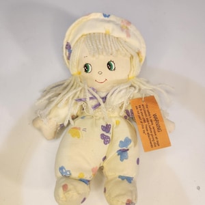 May include: A soft fabric doll with cream-colored yarn hair, wearing a matching bonnet and a cream-colored onesie featuring a colorful butterfly print. An orange rectangular warning tag is attached to the doll's chest with printed text.