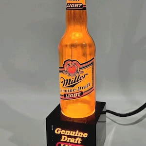 May include: A Miller Genuine Draft Light beer bottle lamp with an orange glow, mounted on a black rectangular base. The bottle features the Miller Genuine Draft Light label, and the base displays the text Genuine Draft LIGHT in illuminated letters.