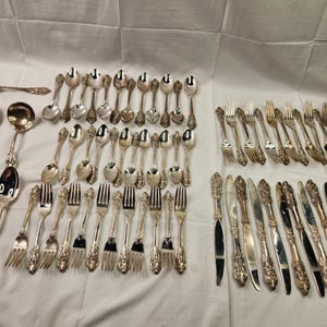 May include: A large collection of silver-toned flatware featuring ornate, embossed floral detailing on the handles. The set includes various sizes of spoons, forks, and knives arranged in rows, showcasing a consistent decorative pattern throughout.