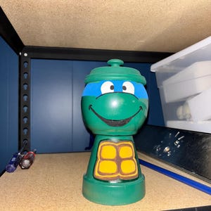 May include: A green painted terracotta pot with a lid, featuring a hand-painted blue eye mask, a black smiling mouth, and a yellow and brown turtle shell design on the front, resembling a cartoon turtle character with a rounded head and body.