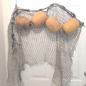 May include: A weathered fishing net featuring four cylindrical, tan-colored foam floats attached to a thick, dark rope along the top edge. The netting is made of thin, light-colored cordage with a diamond-shaped pattern throughout the structure.