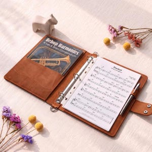 May include: A brown leather binder with a metal ring mechanism, holding sheet music titled Stardust. The left pocket contains a book titled Bebop Harmonies: A Sax & Brass Compendium. A silver pen is clipped to the side, and a small figurine sits nearby.
