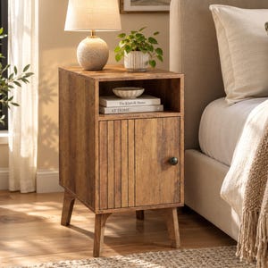 May include: A wooden nightstand with a light brown wood grain finish, featuring an open top shelf and a lower cabinet door with vertical slats and a small round knob. The shelf holds two books titled The Kinfolk Home and AT HOME.