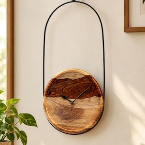 May include: A circular wall clock featuring a natural wood grain face with a distinct dark brown upper section and a lighter brown lower section. The clock is suspended by a thin black metal frame that loops upward to hang from a wooden wall hook.