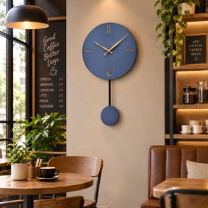 May include: A circular blue wall clock with gold-coloured hands and markers. The clock features a vertical black rod extending downwards to a matching blue circular pendulum. The design includes simple gold lines for the twelve, three, six, and nine o'clock positions, providing a clear and functional timekeeping display.