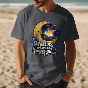 Może przedstawiać: Ciemnoszary t-shirt z krótkim rękawem z grafiką uśmiechniętego żółtego półksiężyca czytającego otwartą książkę ze świecącymi stronami. Napis "Meet me where the pages glow" jest wydrukowany stylizowaną niebieską czcionką pod księżycem i gwiazdami.