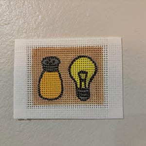 May include: A rectangular piece of white plastic canvas featuring a needlepoint design on a tan background. The design depicts a yellow salt shaker with a gray top next to a yellow light bulb with a gray base, both outlined in black thread.