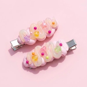 Puede incluir: Dos pinzas para el cabello con una base rosa claro y una cobertura ondulada de color crema translúcido. Las pinzas están decoradas con pequeños dijes de plástico de colores, incluyendo estrellas, corazones y esferas en tonos amarillo, rosa y verde.